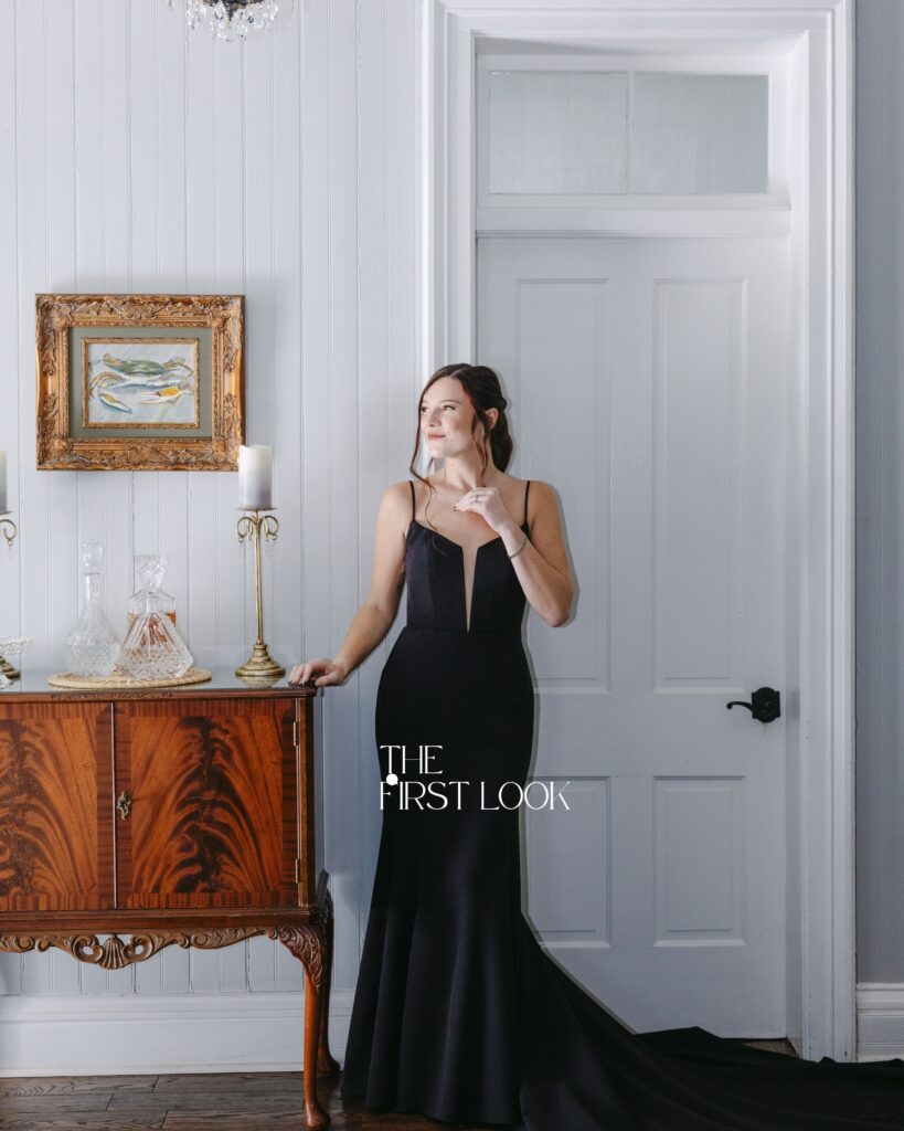 A bride getting ready in a black wedding gown for her first look standing in a white hallway with antique furniture surrounding her