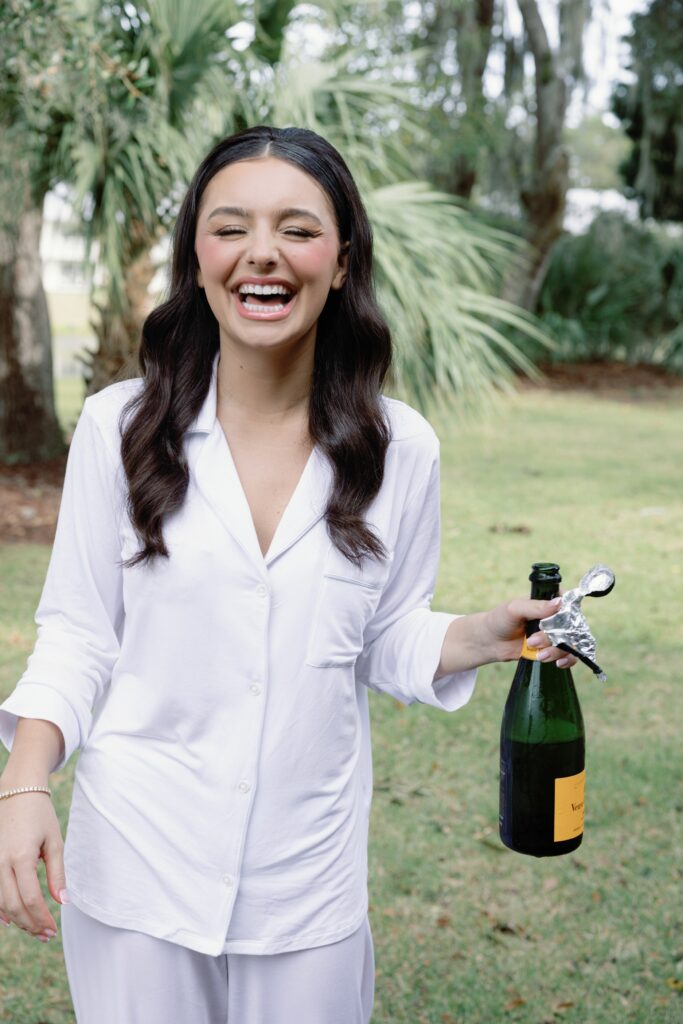 Bride in white pajamas on her wedding day popping a bottle of champagne and laughing