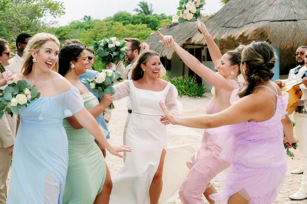 Super happy bride dancing with her bridesmaids that are wearing colorful dresses after she was married with their hands and flowers in the air