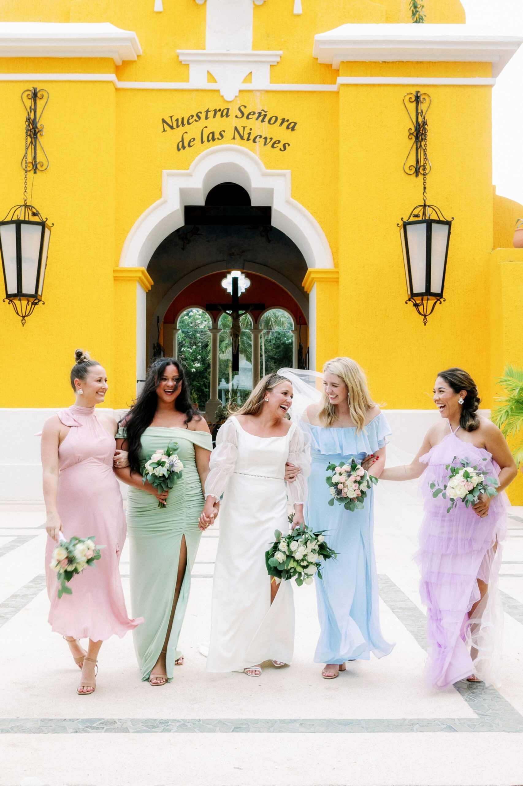 Bride and bridal party celebrating after the wedding wearing pastel colors in front of a yellow chapel laughing