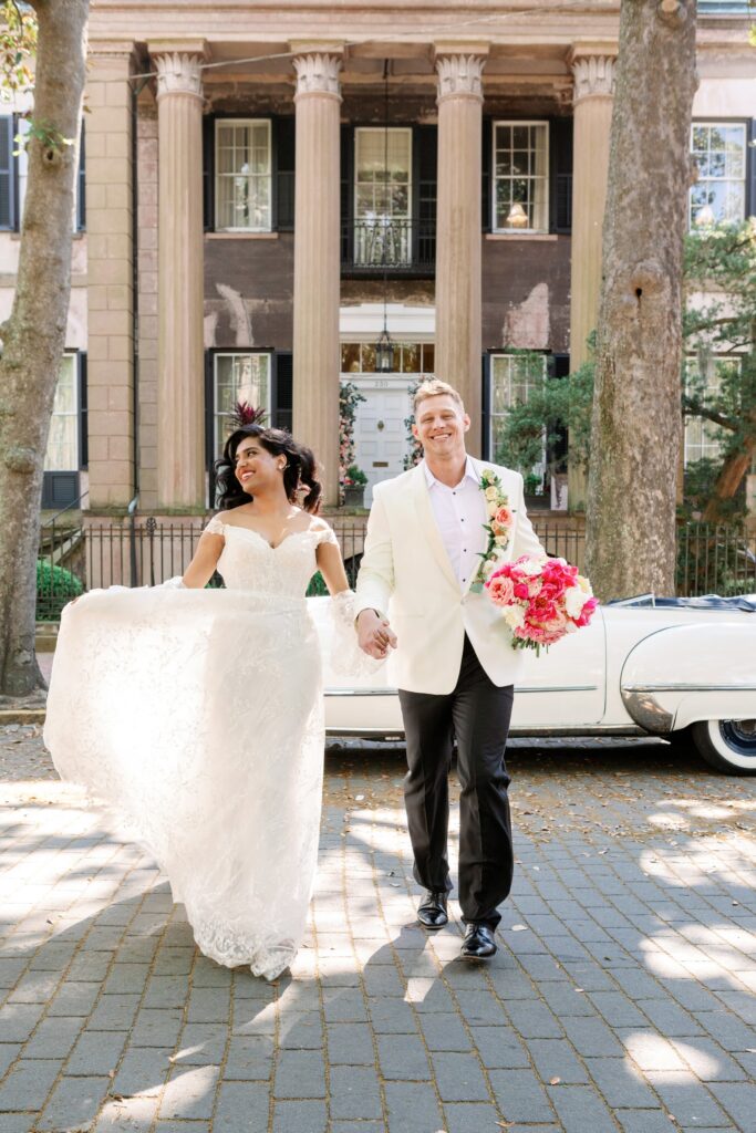 Bride and groom in Savannah, Ga running across the street in front of a historical building with a antique convertible in the background