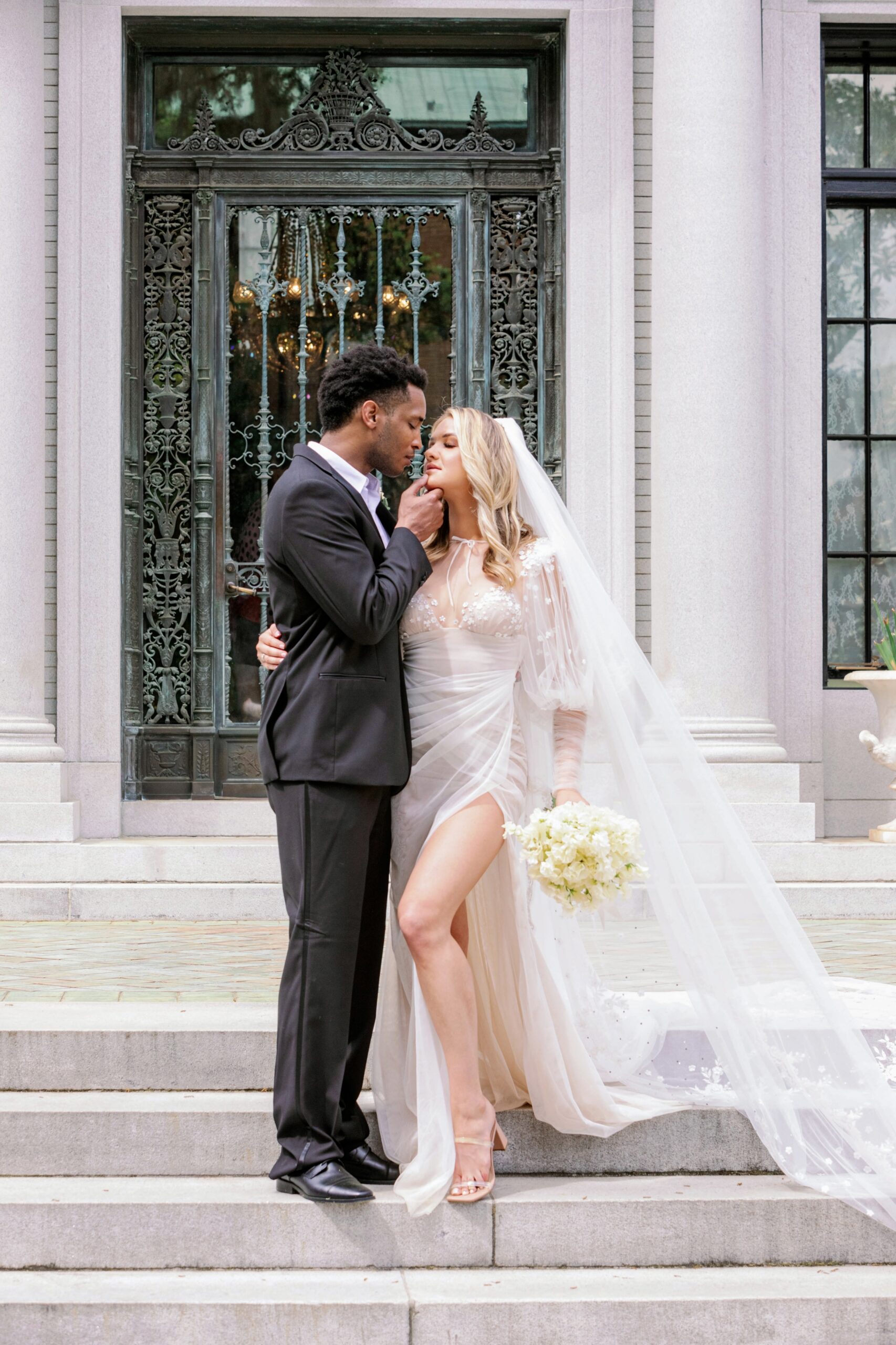 A couple that has just gotten married standing outside the Armstrong Kessler mansion on the steps with him about to kiss her while she holds her flowers