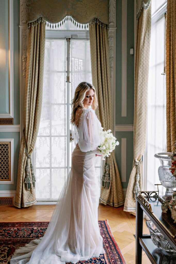 A gorgeous bride in a gown with a veil looking out the window in a french inspired room with velvet curtains at the Armstrong Kessler Mansion in Savannah